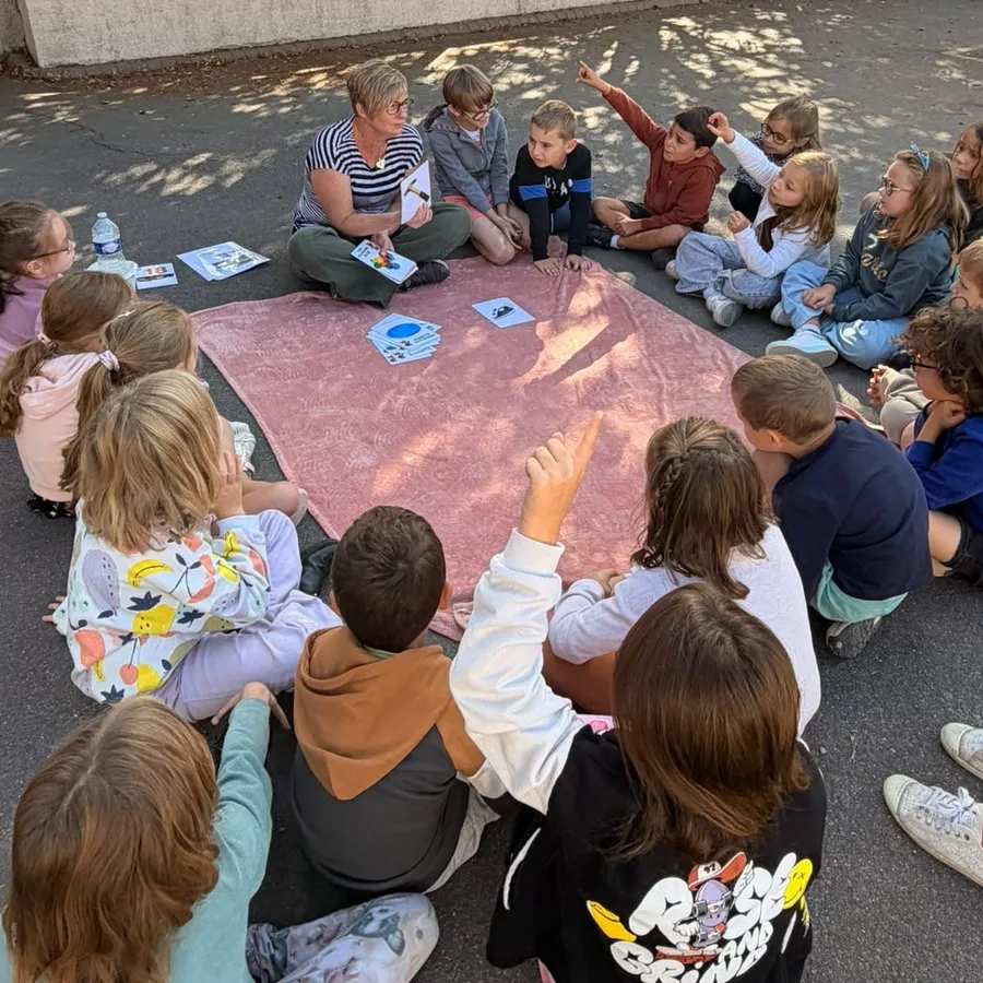 Cécile Tiscornia assise en cercle avec une vingtaine d'enfants, sur une couverture posée dans une cour d'école