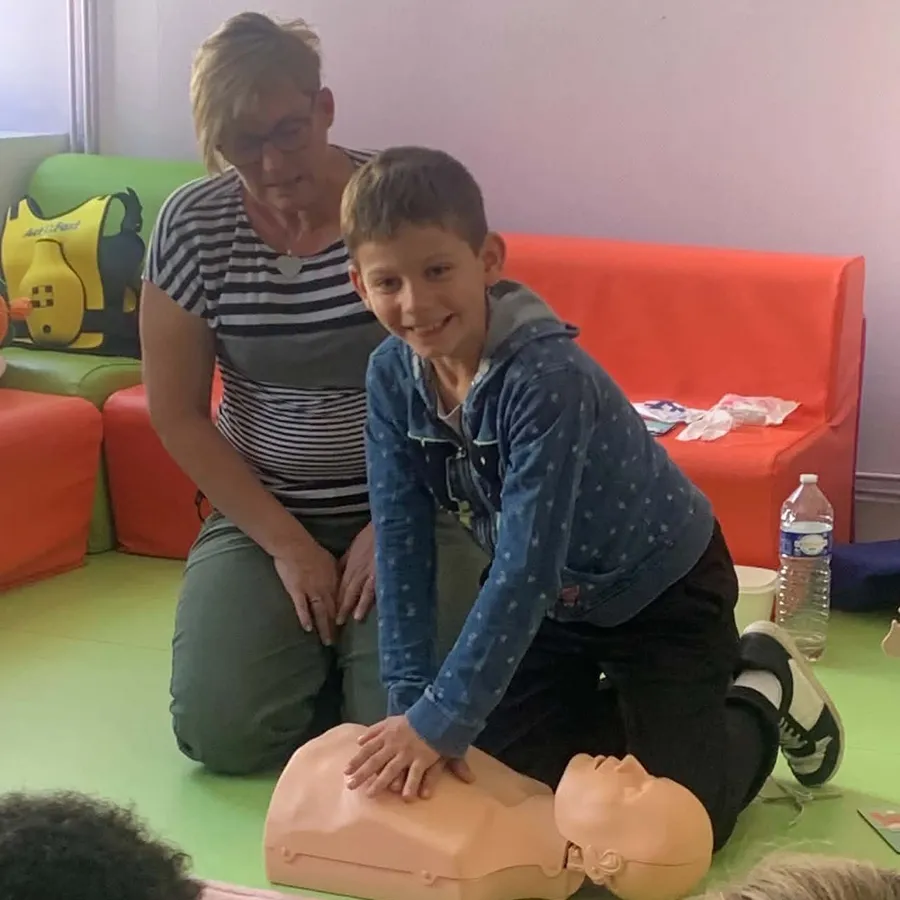 Un enfant réalise des compressions thoraciques sur un mannequin adulte, guidé par Cécile Tiscornia agenouillée derrière lui