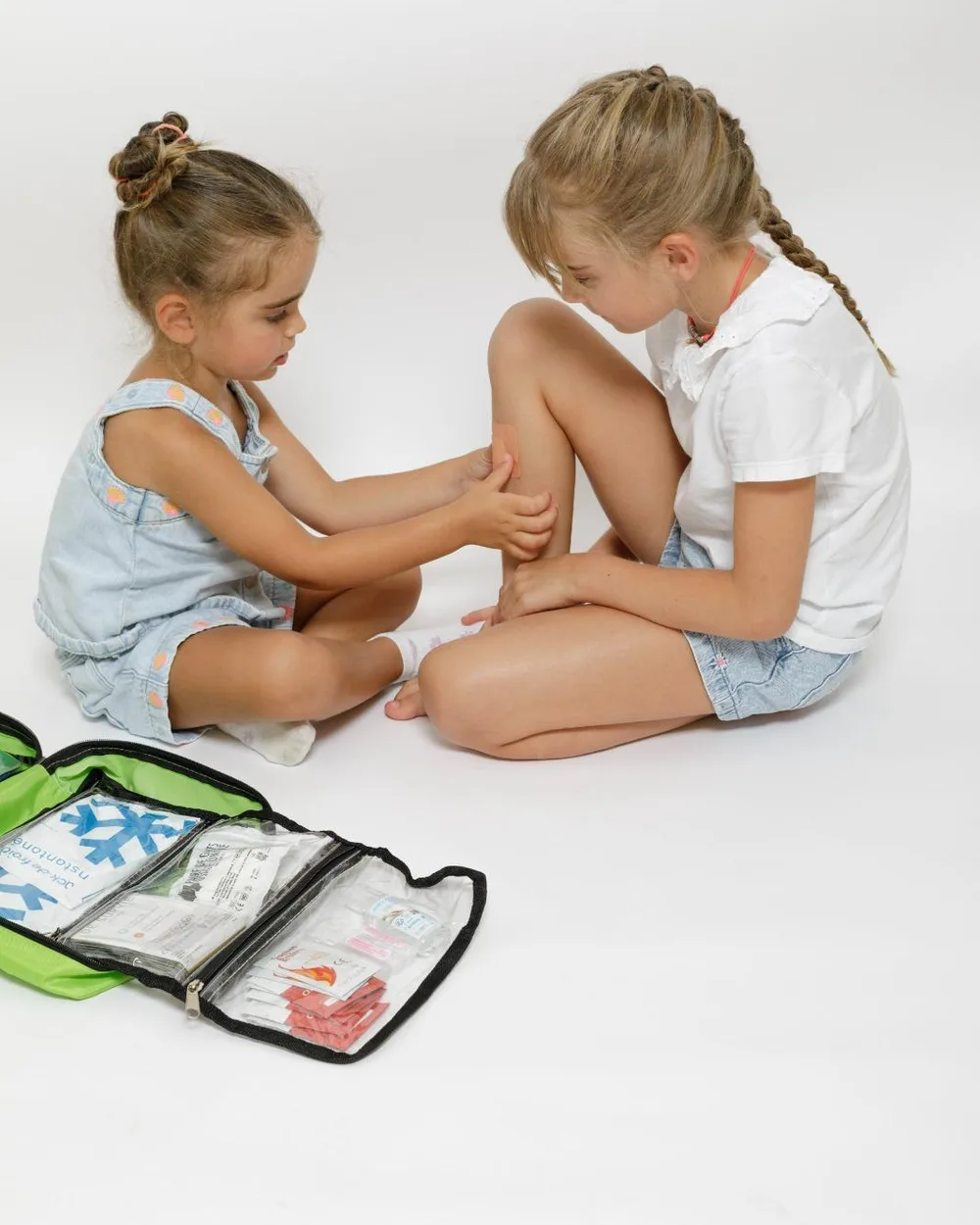La trousse de secours Famille de Sauveteurs ouverte, compartiments transparents visibles, pendant qu'une enfant pose un pansement sur le genou d'une autre